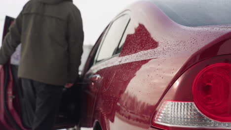 rear side view of red car covered with water droplets as businessman in winter jacket opens door and enters to drive, with surrounding parked cars reflected on glossy surface in snowy outdoor setting