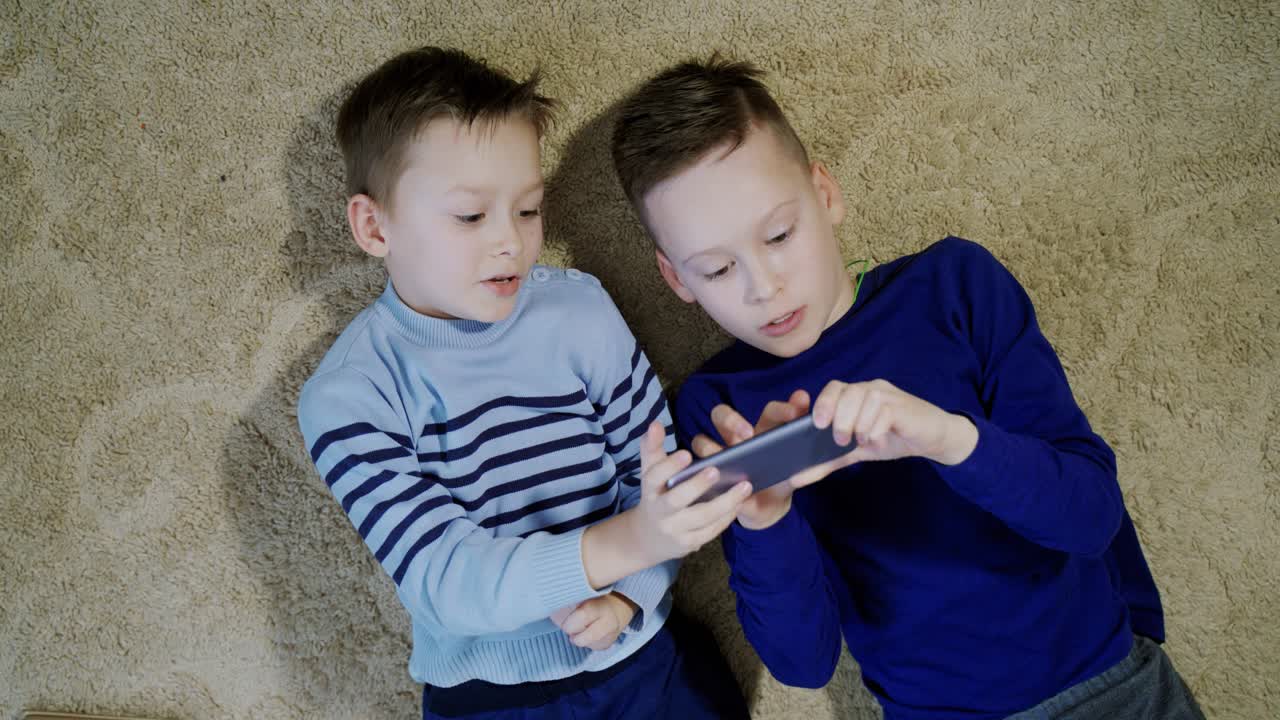 Two happy boys lying on the floor and playing on one smartphone together. Relaxed kids having fun altogether with modern gadget on a soft rug. Close-up