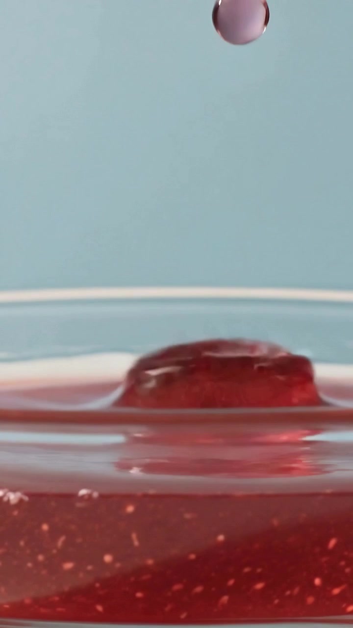 Video verticale: Versamento di sciroppo rosso che rivela bolle e forma una goccia in un piatto di vetro poco profondo.