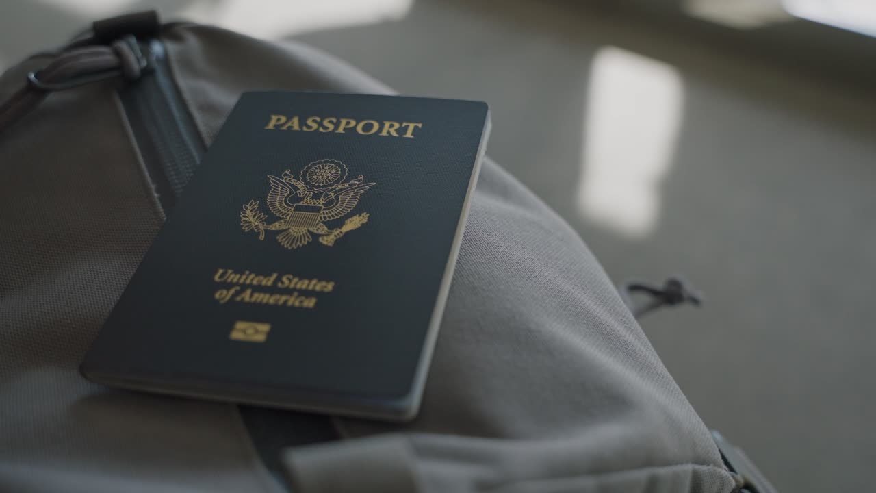American passport on backpack in airport terminal with shadow and silhouette of passengers walking by boarding plane or passing with luggage and suitcases in daytime by lounge or gate sitting area