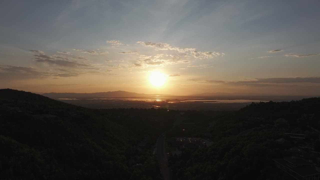Aerial drone dolly out tilting down shot from Bountiful Canyon in Utah at golden hour with road, forest, valley homes, Wasatch Mountains, cityscape, and the Great Salt Lake at sunset