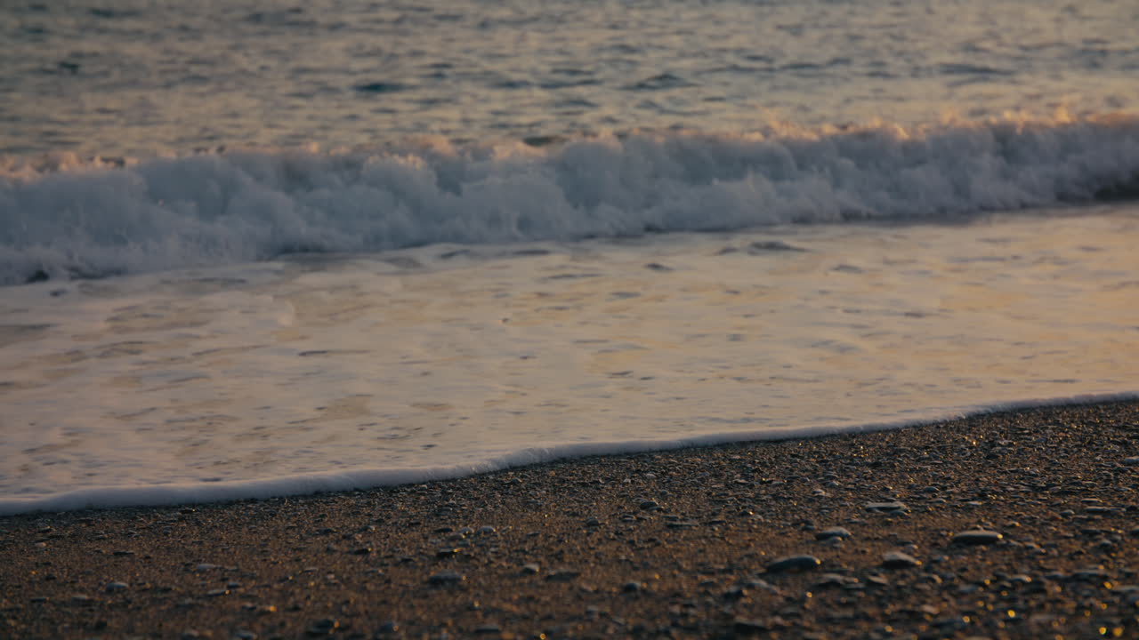 Low-angle view of waves softly reaching the pebbled shore in warm sunset light. Peaceful and detailed coastal atmosphere