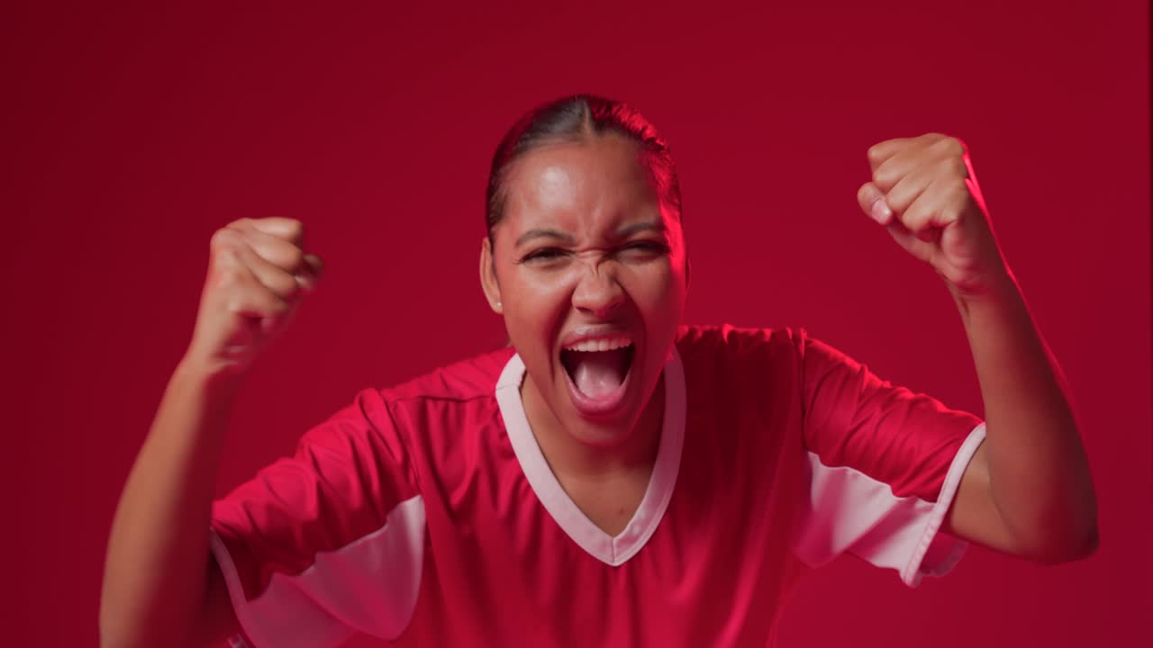 African woman shouting and pumping fists while tugging red-and-white jersey in studio after goal