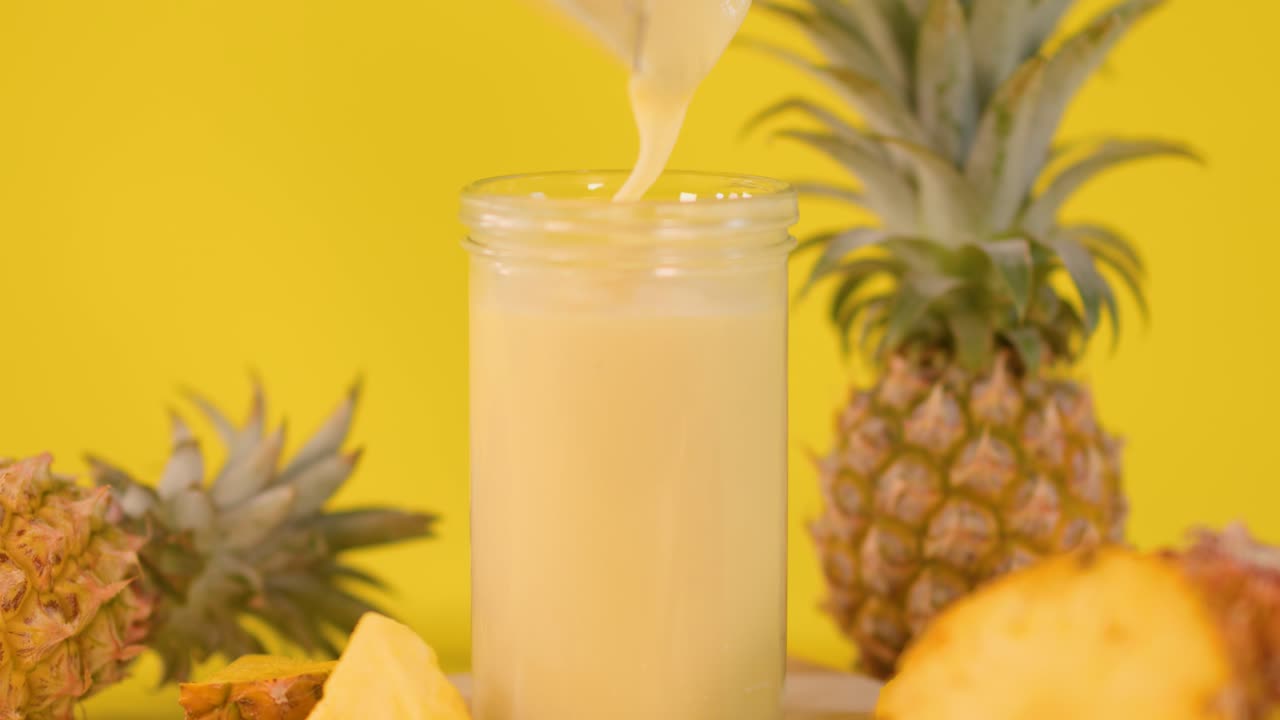 Creamy pineapple smoothie poured into glass, surrounded by fresh fruit, bright yellow background, static shot
