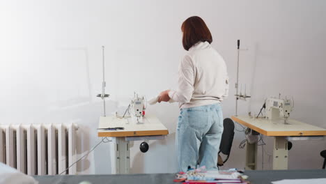 outfitter walks to sewing machine carrying scissors and white fabric from large table filled with fabrics, sits on sewing chair, places fabric on needle to start sewing process