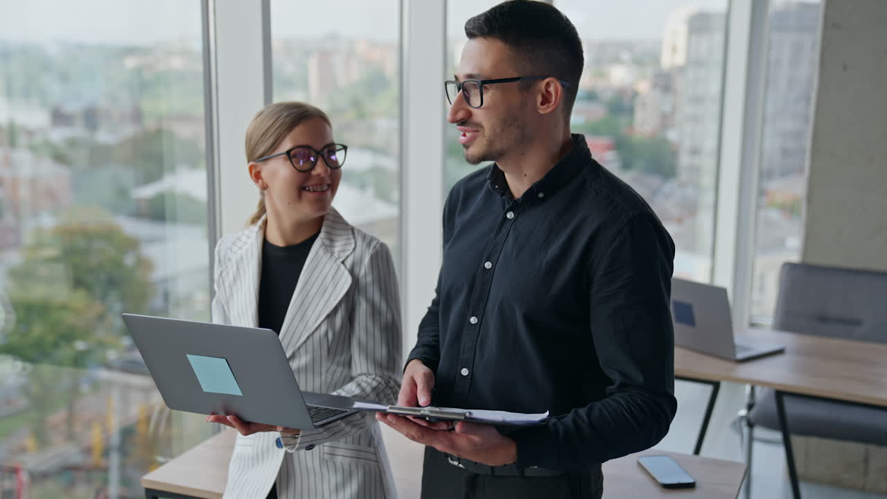 Friendly talking colleagues standing at the desk in office. Male and female employees holding papers and laptop speaking and smiling.