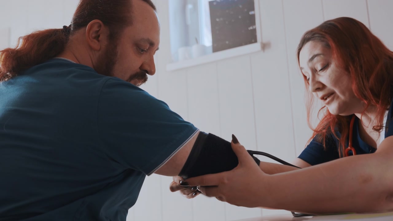 Community nurse measuring blood pressure white patient in bright home office near window, calm conversation, stethoscope ready, manual cuff positioning, focused measurement, natural light