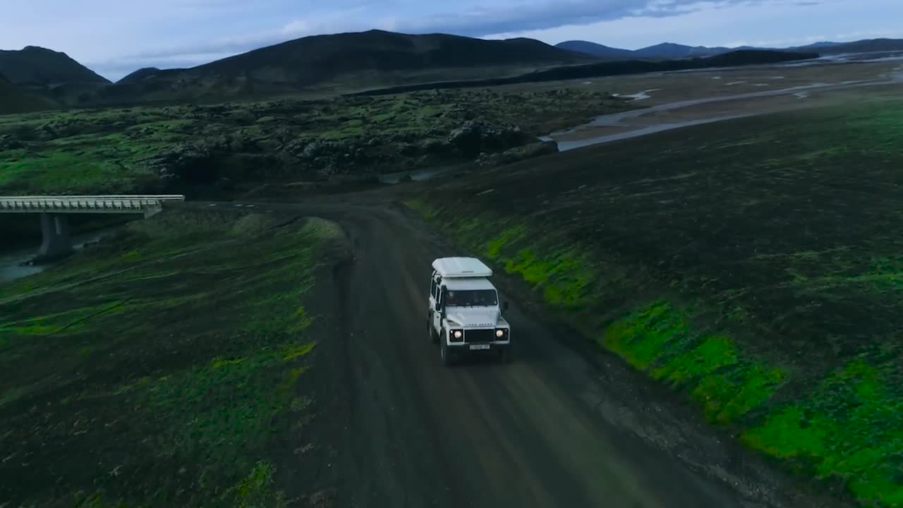 Aerial drone gliding and flying over a 4x4 white Jeep or truck that is driving on a wet and damp muddy road during evening or dusk in Iceland or Greenland. A foggy river and bridge in the background.