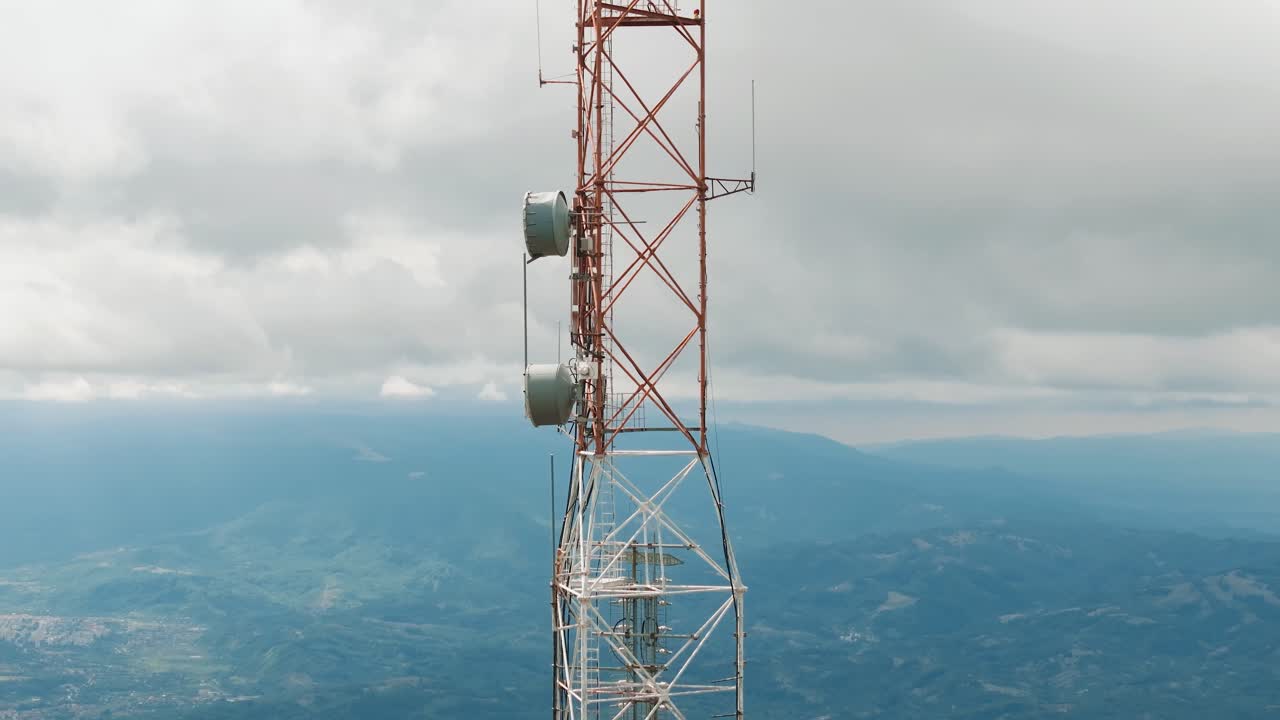 High altitude telecommunication tower holding multiple antennas, over a mountain landscape with distant small villages, aerial view