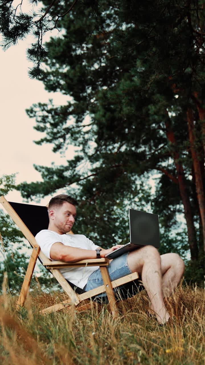 Approaching a man sitting in the folding chair. Freelancer works on laptop remote. Low angle view. Nature at backdrop. Vertical video