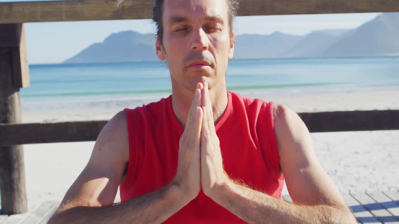 Video di un tranquillo uomo caucasico con dreadlocks che pratica meditazione yoga seduto in spiaggia sotto il sole