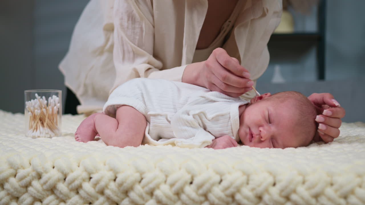 Madre pulisce delicatamente l'orecchio di un neonato addormentato con un batuffolo di cotone su una morbida coperta. Igiene e cura della madre.