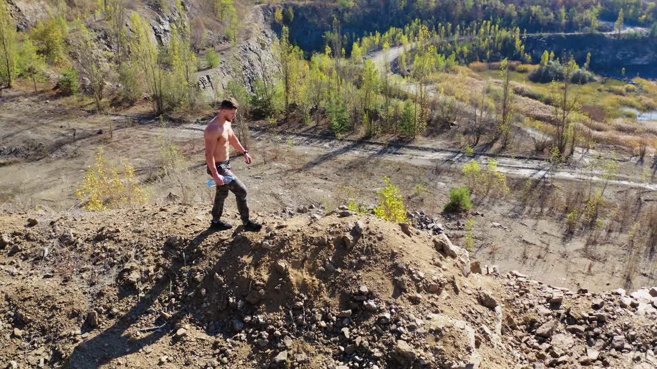 Young sportsman with a bottle of water in nature. Shirtless athlete drinking water after the workout in the mountainous area. Aerial view.