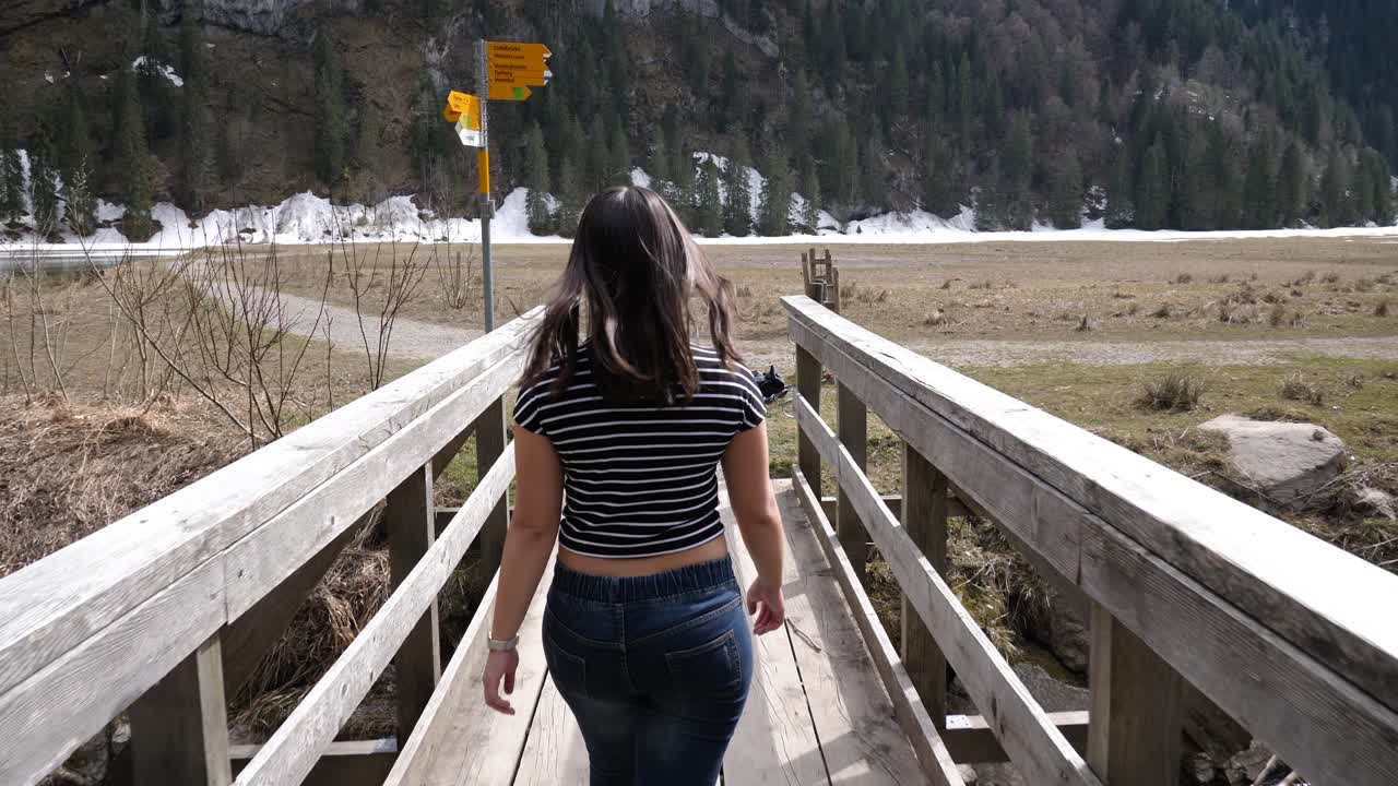 joven asiática con cabello largo y camisa a rayas y jeans camina sobre un puente de madera en suiza en cámara lenta