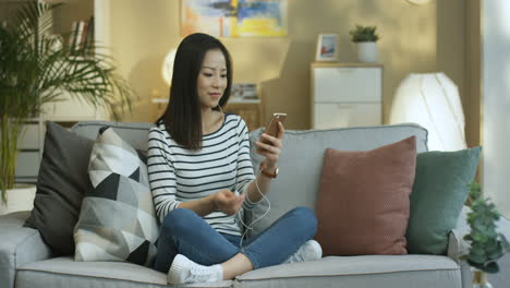 Mujer-Joven-Con-Camisa-A-Rayas-Y-Auriculares-Escuchando-Música-En-Un-Smartphone-Sentada-En-Un-Sofá-En-La-Acogedora-Sala-De-Estar