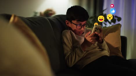 Kleiner-Junge,-Der-Abends-Zu-Hause-Auf-Dem-Sofa-Sitzt-Und-Auf-Sein-Mobiltelefon-Schaut,-Auf-Dem-Emojis-Mit-Bewegten-Grafiken-Zu-Sehen-Sind,-Die-Mehrere-Benachrichtigungen-über-Soziale-Medien-Oder-Messaging-Dienste-Zeigen,-Online-Inhalte-Liken-Und-Darauf-Reagieren