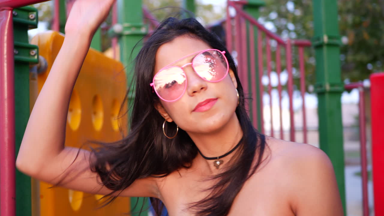 A young woman hipster wearing vintage fashion clothing and retro pink aviator sunglasses and smiling in a park playground SLOW MOTION