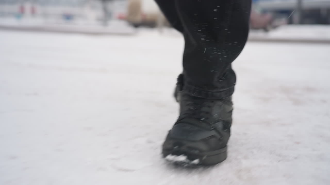 Close up lower angle view of person wearing black boots walking across snow covered ground with snowflakes falling and parked cars visible as he enter a car in background during cold winter day