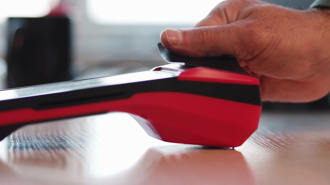 Tight low angle view shows red and black handheld device resting on wood tabletop while human hand reaches in and grasps upper panel, creating clear hands-on product interaction and pickup moment