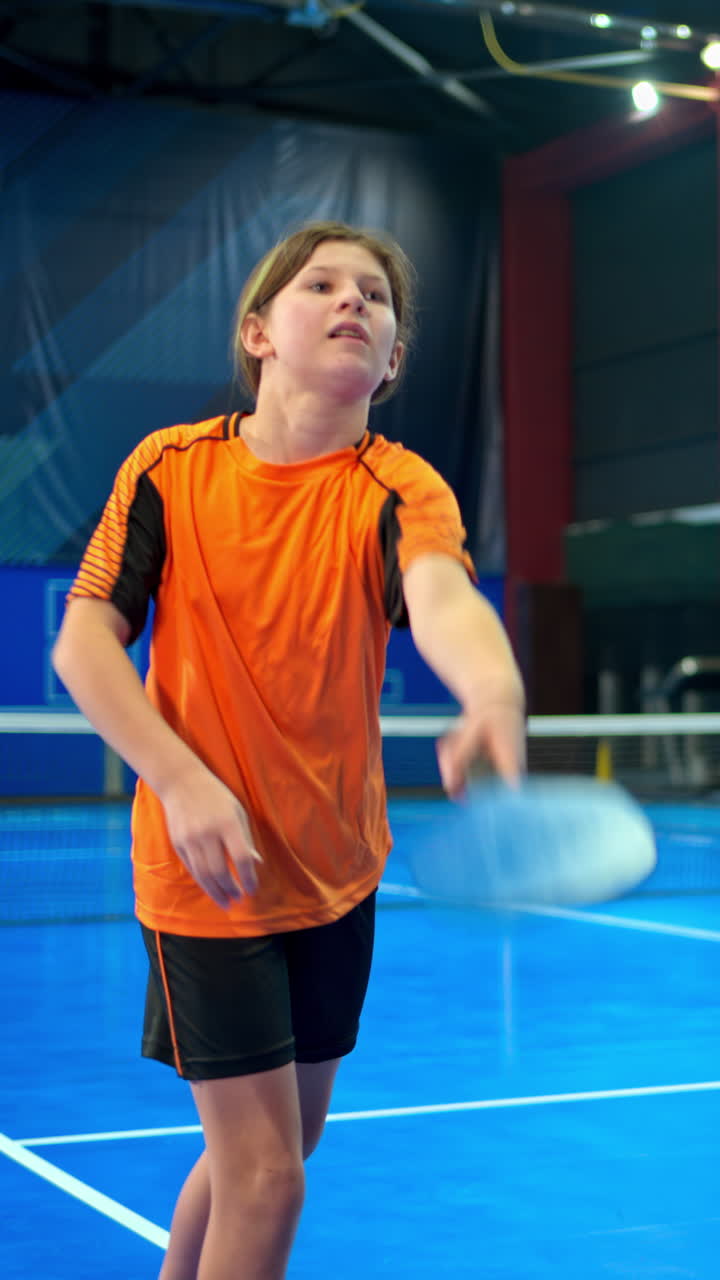 A woman teaching a child to play pickleball on a blue, inside court. Vertical