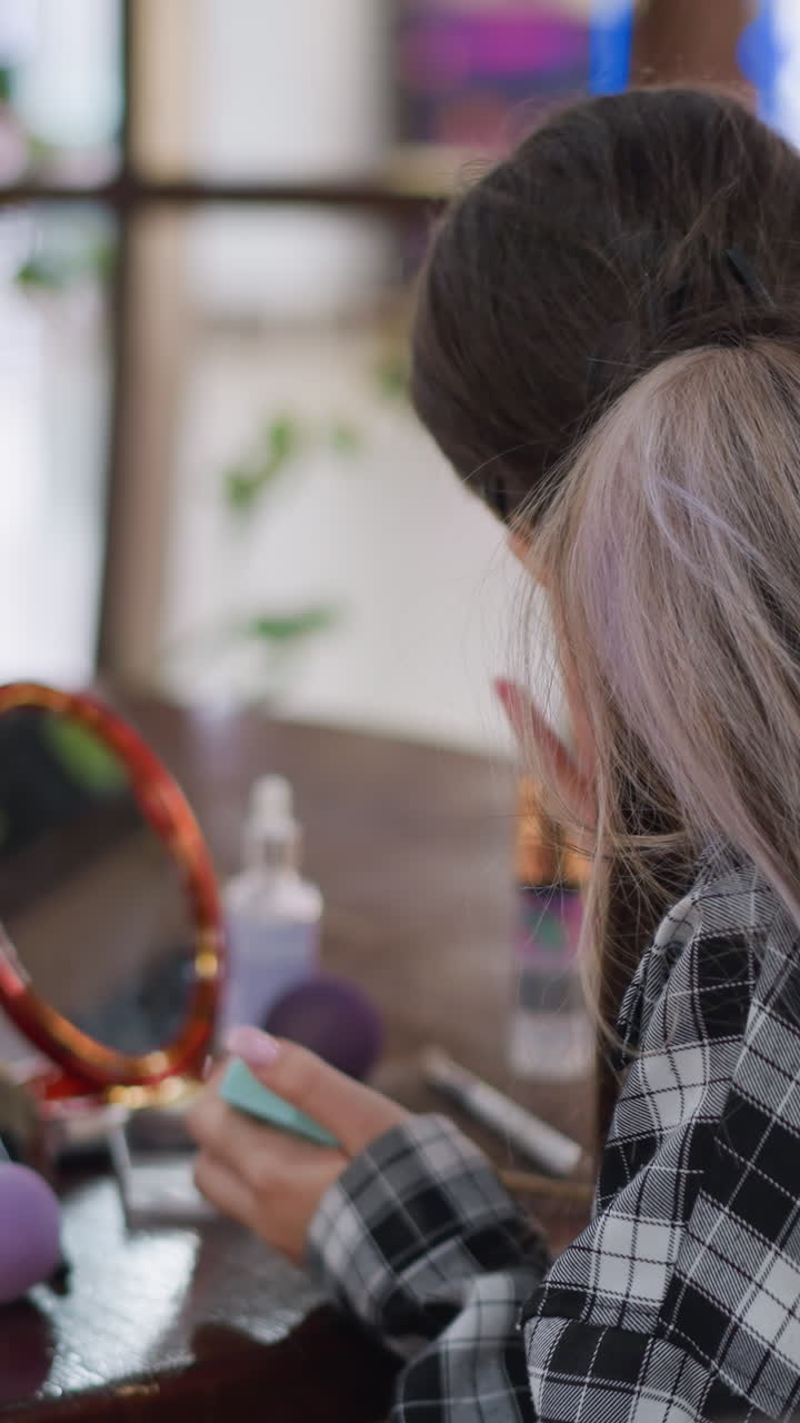 Youth beautifies herself, Teen in mirror attending grooming, Teenager applies beauty regimen in softly lit room, Young girl prepares herself using various grooming tools in gentle natural light