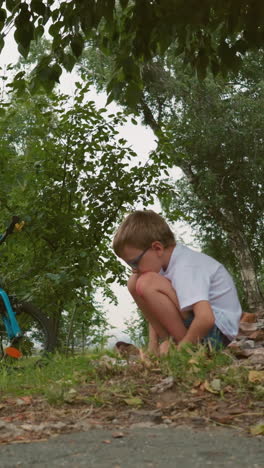 un niño se agacha bajo la sombra de un gran árbol, con una bicicleta negra estacionada cerca, el niño parece tranquilo y reflexivo, rodeado de vegetación