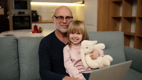 grandfather and small granddaughter sitting on sofa and looking to the camera