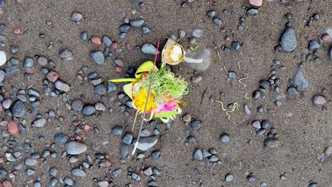 top down of balinese hindu canang sari offering to gods and spirits with food, flowers, and smoking incense stick on beach on tropical island of bali indonesia