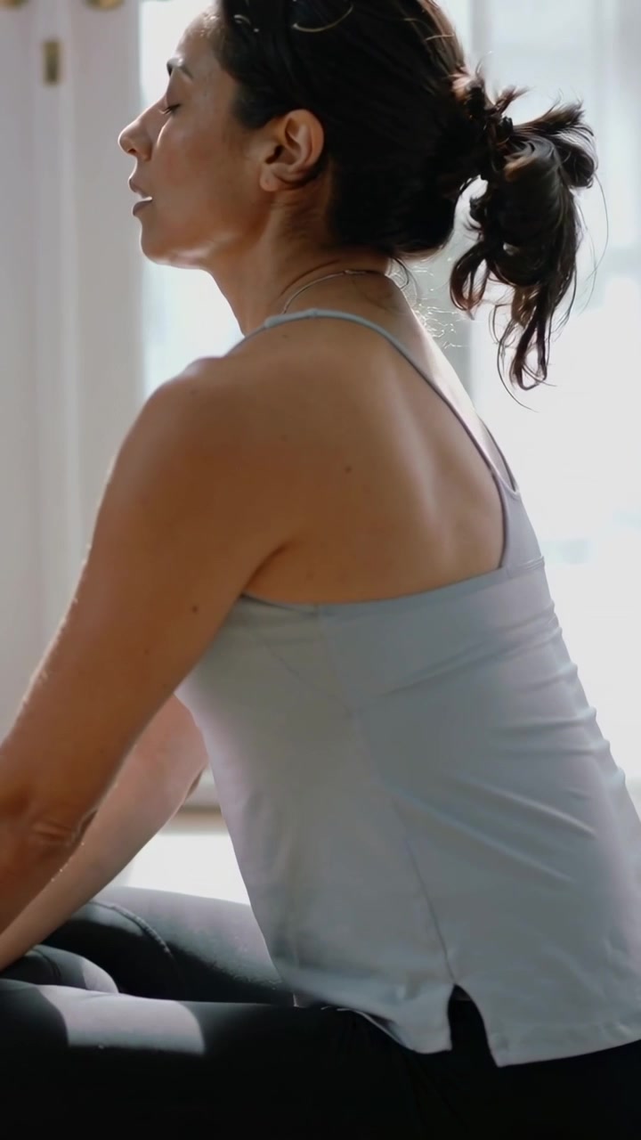 Vertical video: Woman breathing by glass door with sun falling, calming in blue top and black pants