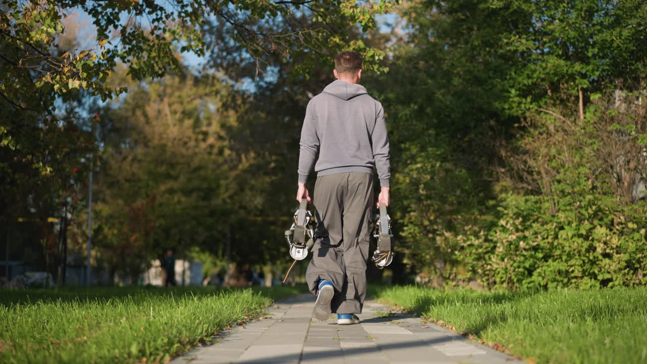 緑の木や草に囲まれて,明るい自然の光の下で,グレーのフーディ,グレイのスウェットパンツ,青いスニーカーを履いて,公園の舗装された道を歩いている男性
