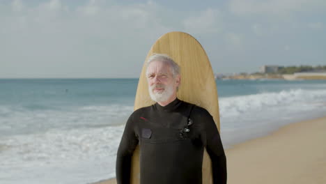 front view of a smiling senior man standing on seashore with surfboard and looking at the camera