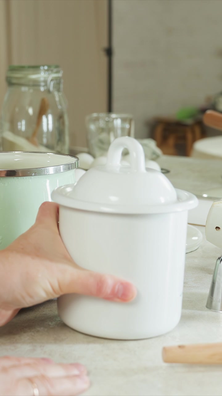 Hand with ring on ring finger interacts. Countertop features glass jar. Person in green garment is visible in background handling white lid and green pot Scene is set
