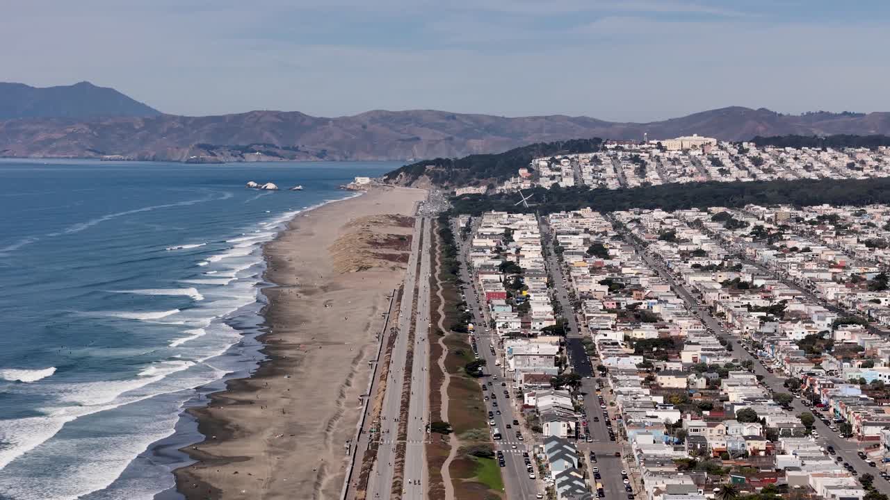 Drone view of Ocean Beach with rolling waves, wide sandy shore, and the Great Highway running alongside residential buildings and dunes.