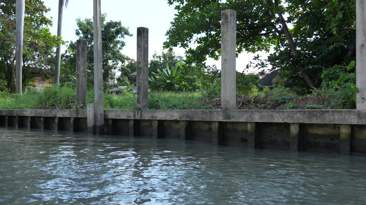 Low-angle boat perspective of a concrete canal embankment lined with tall utility poles and thick vegetation, typical of Bangkok’s klongs and riverside districts with buildings and local greenery