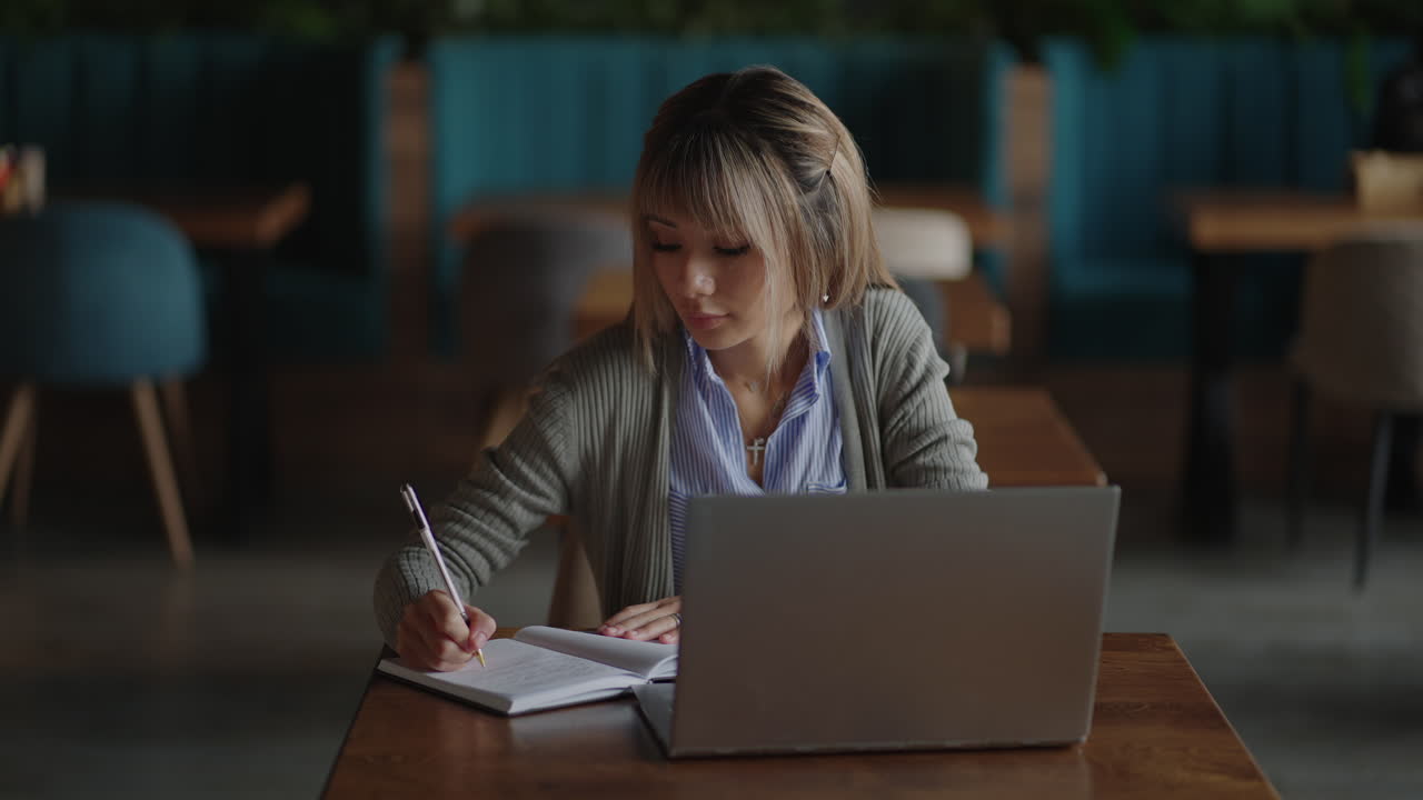Asian woman working on her laptop and writing in his notebook sitting at a table. Working in coffee shop. woman looking to a laptop screen and making notes in her notebook. studying online.