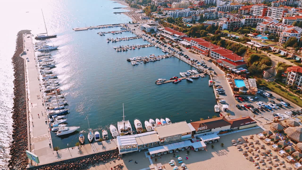 Big and small boats stand at the berths of the yacht club. Multiple cars are parked nearby. Sunny resort of Sveti Vlas, Bulgaria. Aerial view