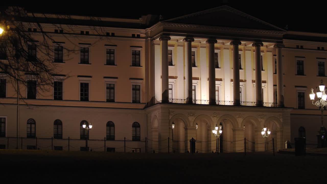 Cinematic slow motion wide 4K shot with parallax motion of tree branches in front of illuminated front and entrance of the Norwegian Royal Palace on top of Karl Johan street, at night in Oslo Norway