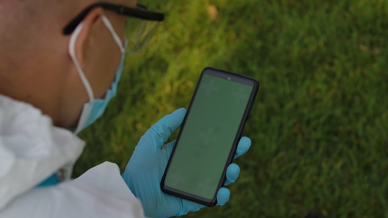 una vista detrás del hombro de un hombre con traje de protección sosteniendo un teléfono inteligente con una pantalla verde con una tecla de croma, navegando por el contenido sin tocar o deslizar. cerrar. cámara lenta