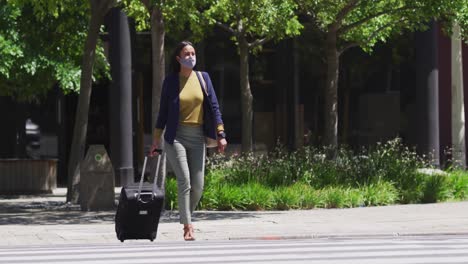 African-american-woman-wearing-face-mask-crossing-road-wheeling-suitcase