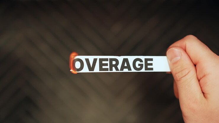 Close-up of a hand holding a paper label reading “COVERAGE” as the edge burns with a flame on dark background, symbolizing insurance risk, cancellation, loss, copy space