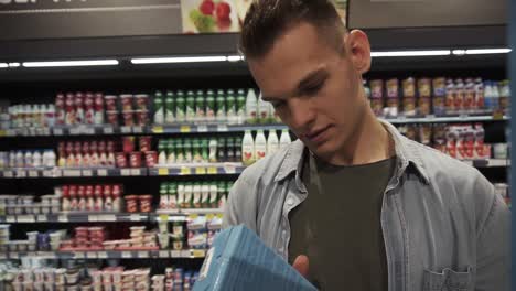 Footage-from-the-store-shelf.-Portrait-of-caucasian-man-taking-a-box-from-a-shelf,-reading-the-label-and-take-it-to-his-cart.-Food-assortment-shelves-on-the-background