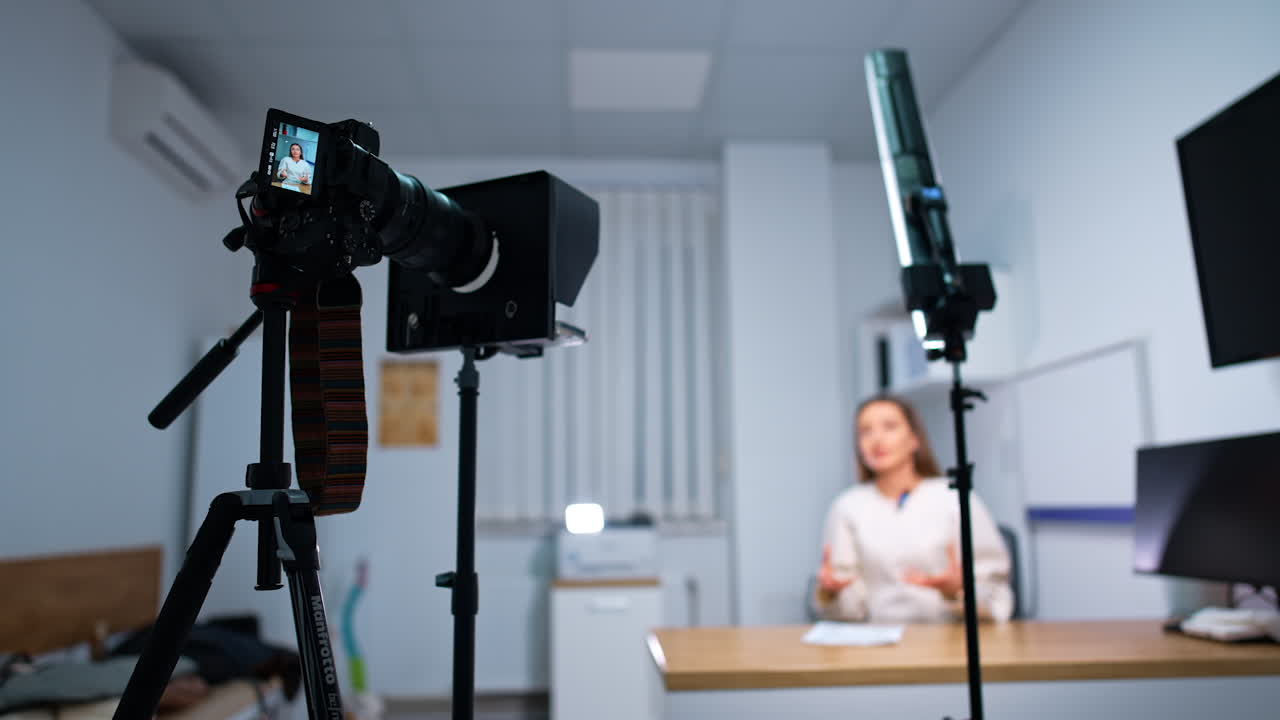 Blogger sits at desk recording a content, speaking to the camera. Professional equipment filming a speaker. Blurred backdrop.