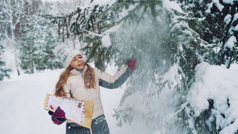 happy child is holding a big envelope and playing with snow. a little girl shakes snow off a fir tree in winter on the snowy background.