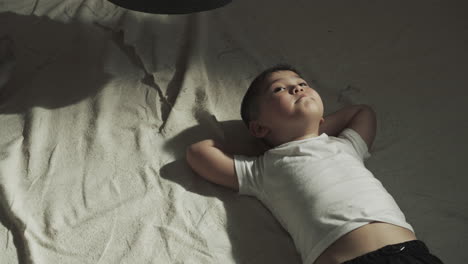 child lies under punching bag in gym after workout. thoughtful little asian boy rests on carpet in martial arts class slow motion. junior athlete relaxes on floor