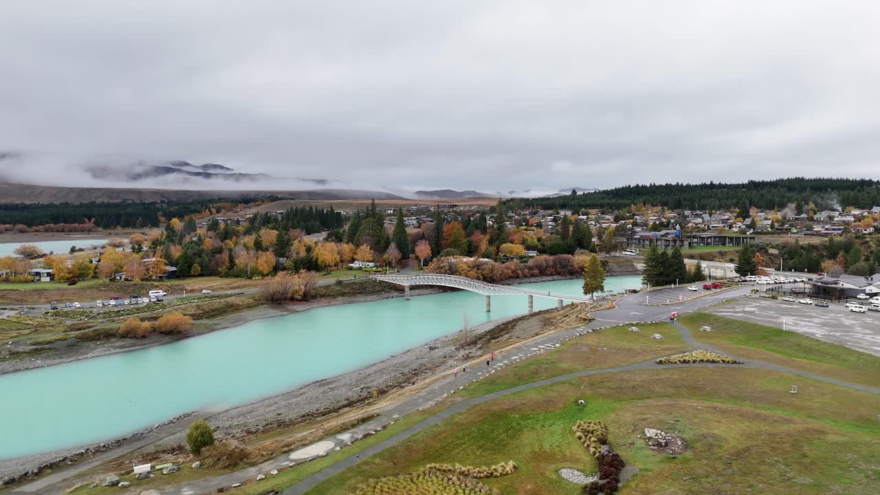 Drone footage captures Lake Tekapo's turquoise waters and autumn foliage under overcast skies, showcasing the serene New Zealand landscape