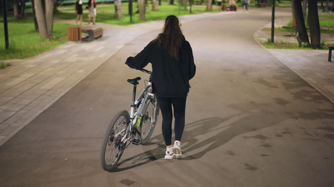 Woman Walking Bike Through Green Park, Casual Jacket And Sneakers, Wooden Benches Lining Path, Tree Canopy, Late Afternoon Light, Relaxed Pace, Urban Oasis Atmosphere, Solitary Stroll
