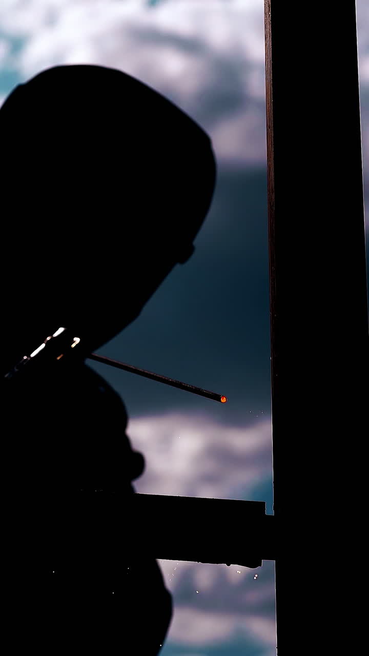 builder in protective wear dark silhouette with operating welding machine at metal fence against sky and clouds