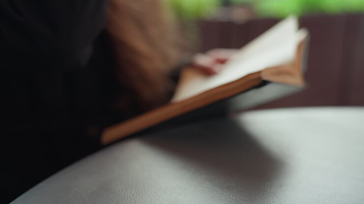 coffee cup and book on table, young woman reading and reaching for cardboard cup in cozy indoor cafe with soft bokeh light, warm steam rising from lid, textured table surface and relaxed study mood