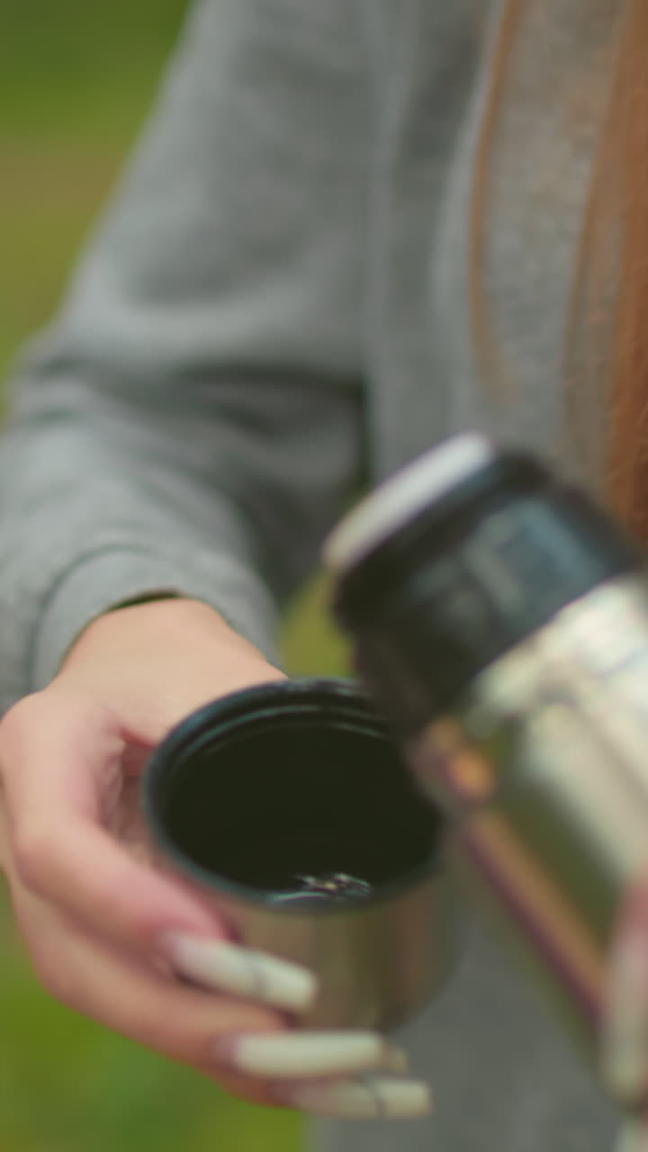 Close-up of young woman in grey sweater carefully unscrewing thermos cap, pressing inner cover to pour water into lid amidst serene green forest, with delicate, well-manicured nails