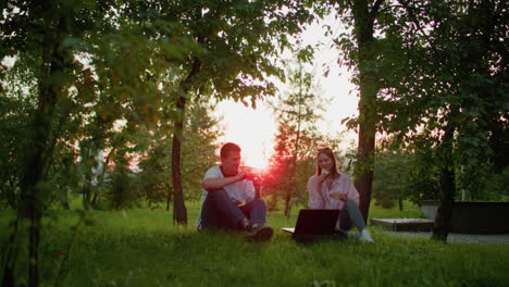 two friends sitting outdoors with a laptop in front of them, toasting with slices of bread, surrounded by lush greenery and trees, enjoying the warm sunlight filtering through the leaves